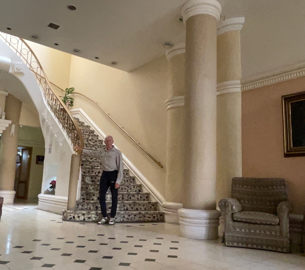 An elderly man stands on the lower level of a spacious foyer, next to an elegant staircase with decorative railing and carpet. Tall columns and a patterned floor are visible, along with a cozy armchair in the background.
