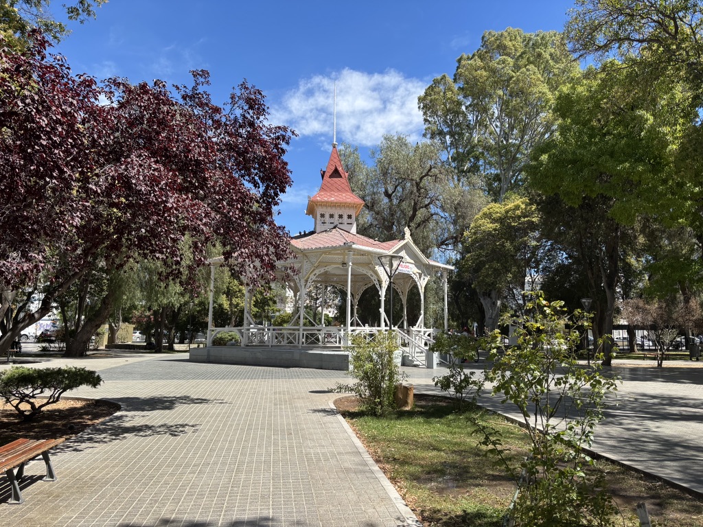 View of a gazebo with a red roof surrounded by trees in a park on a sunny day, with a paved walkway leading towards it.