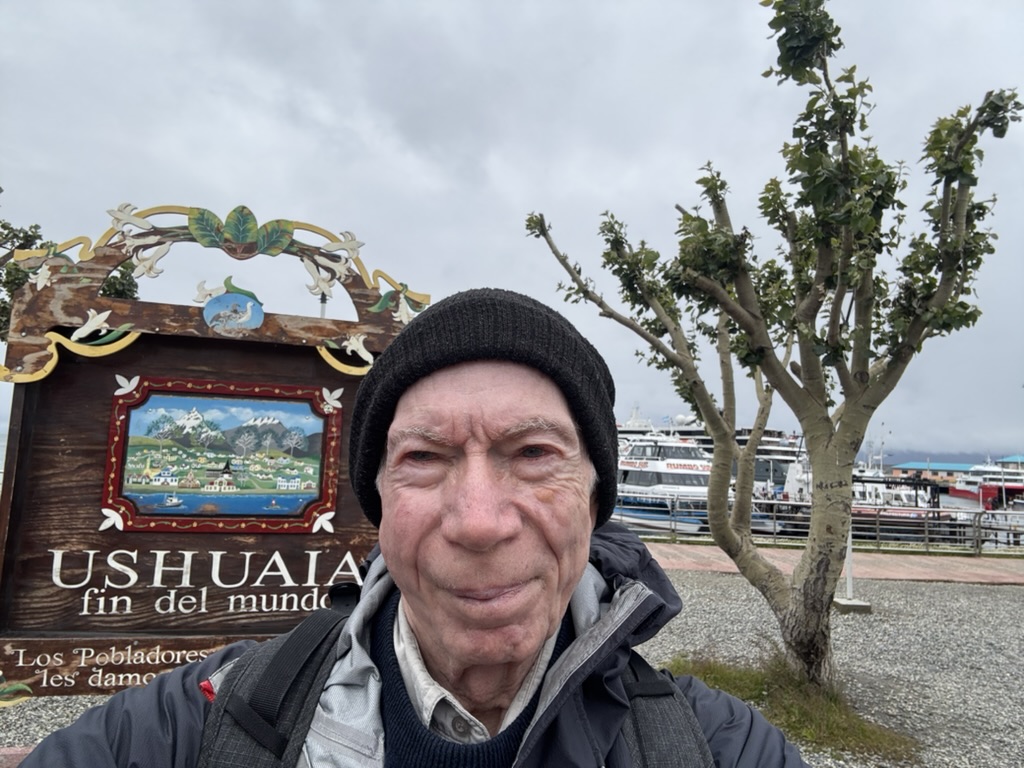 A person wearing a black beanie stands in front of a sign that reads 'Ushuaia, fin del mundo' with a scenic illustration of mountains and houses. The background features a harbor with boats, and a cloudy sky.