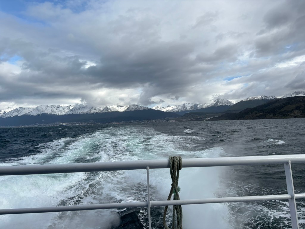 View from a boat showing a mountainous landscape with snow-capped peaks and a cloudy sky, creating a dramatic seascape.
