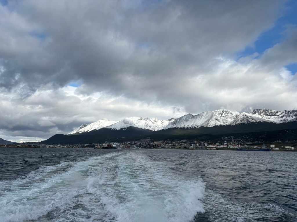A scenic view of snow-capped mountains under a cloudy sky, with a glimpse of a coastal town in the foreground and wake patterns in the water.