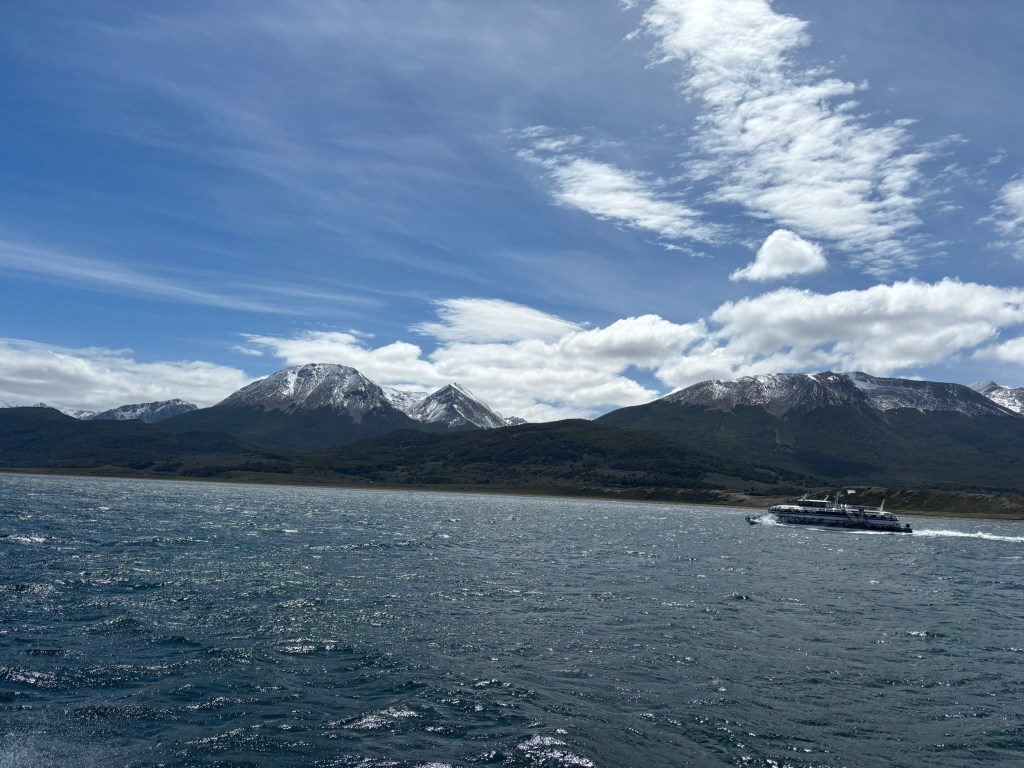 A scenic view of mountains with snow-capped peaks against a blue sky, overlooking a calm body of water with a boat in the foreground.
