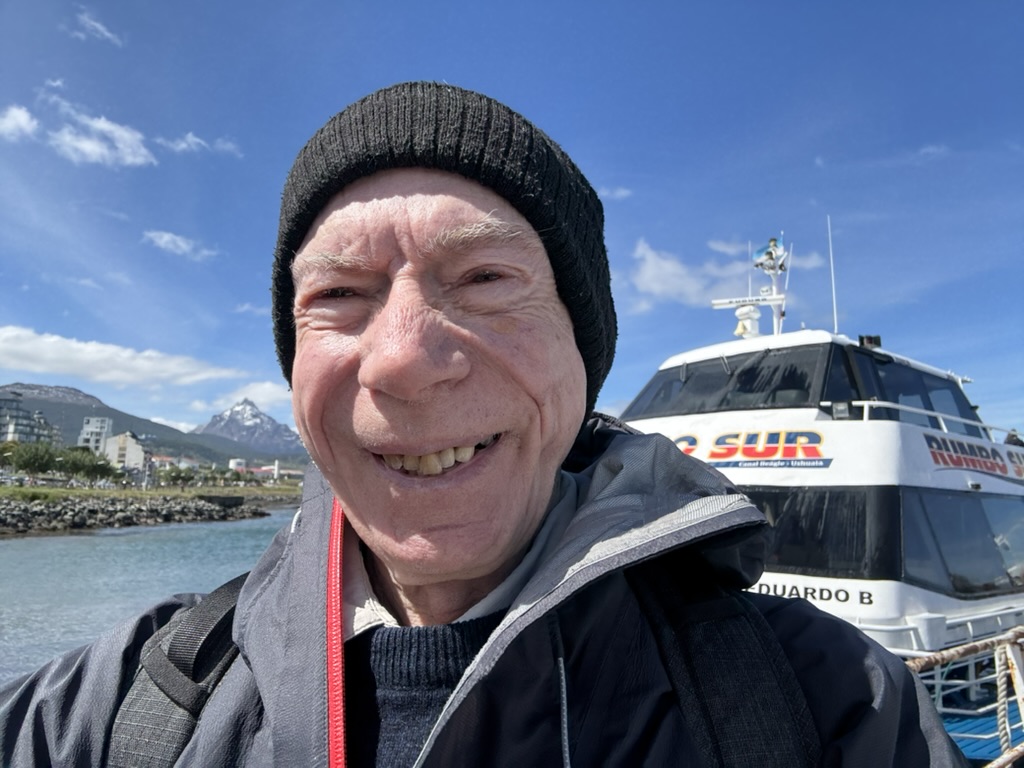 A smiling elderly man wearing a black beanie and a jacket stands in front of a boat with the name 'Eduardo B' visible. The background features a scenic view of mountains and a clear blue sky.