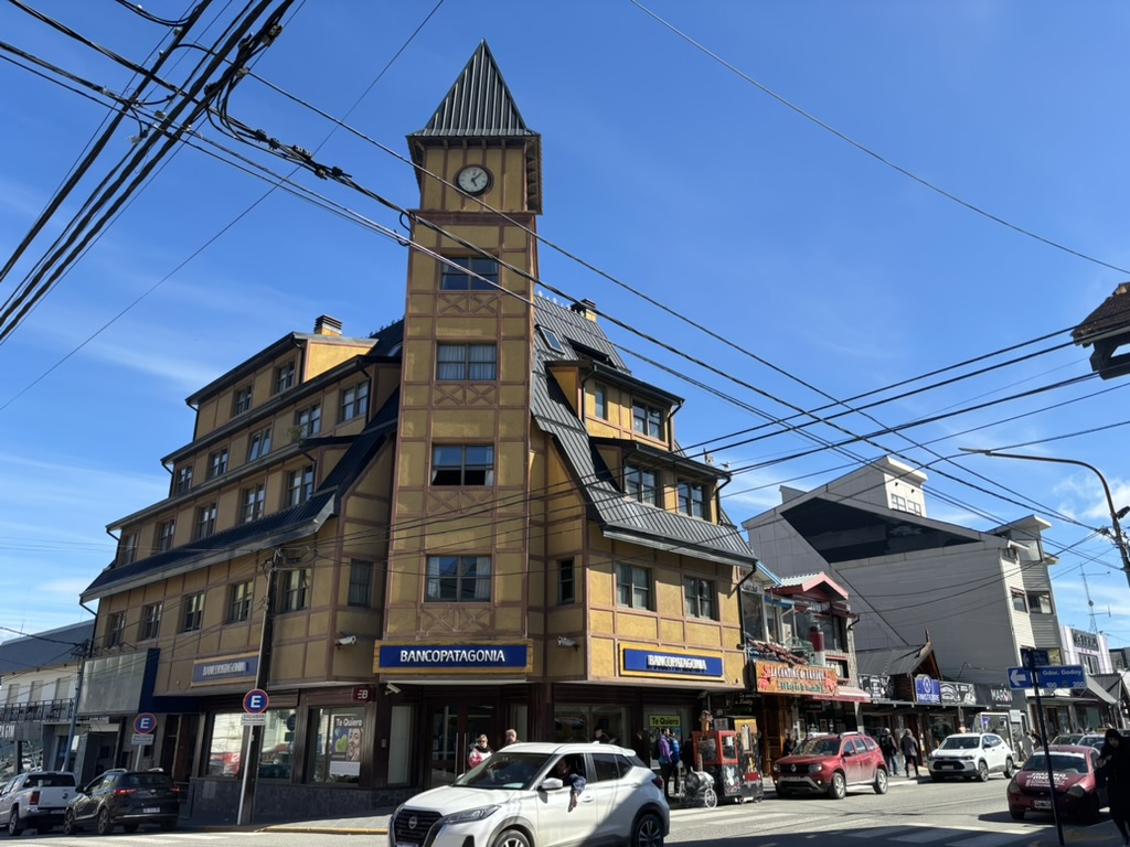 A multi-story building with a clock tower on top, featuring a mix of yellow and brown exterior. The first floor has a bank sign and several shops, with pedestrians walking by and cars parked along the street. The sky is clear with scattered clouds.