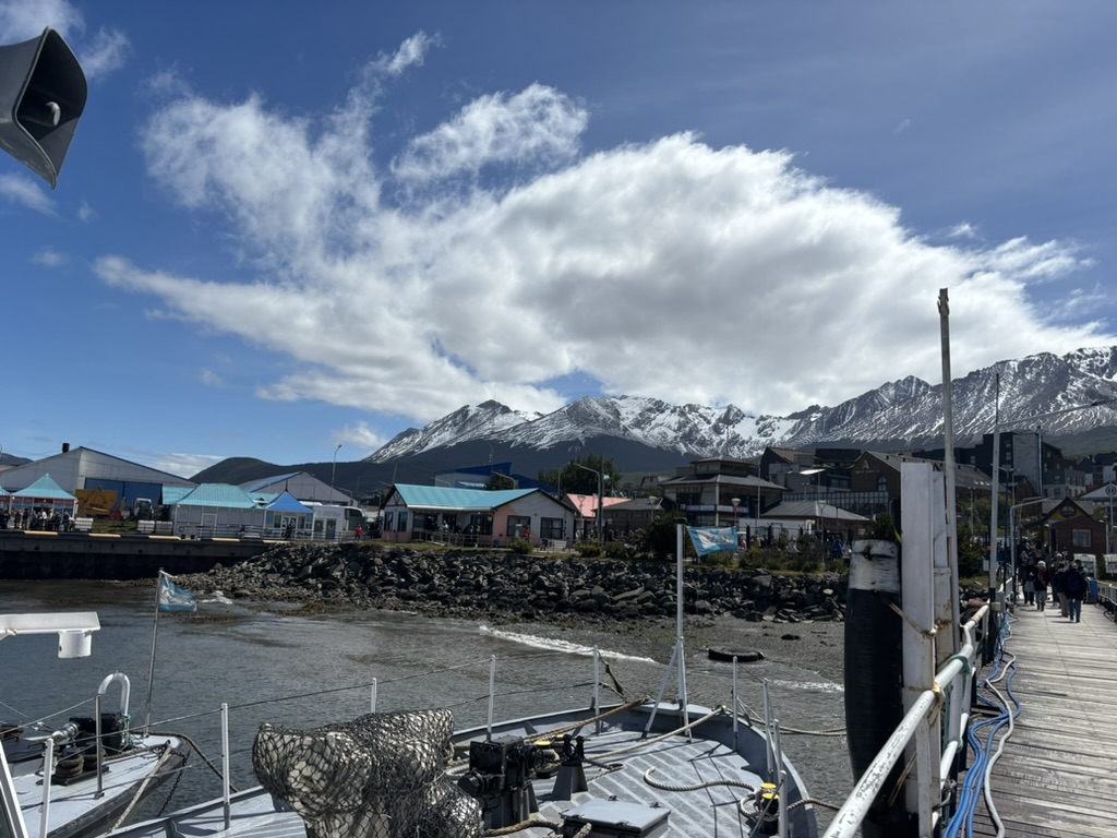 View of a harbor with boats, rocky shore, and colorful buildings, set against snow-capped mountains and a partly cloudy sky.