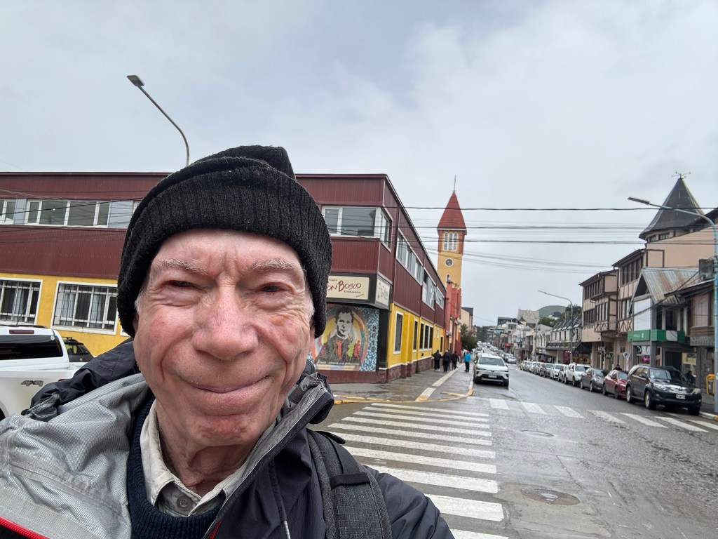 A person smiling in the foreground with a beanie, in a street setting with colorful buildings and a clock tower in the background, on a cloudy day.